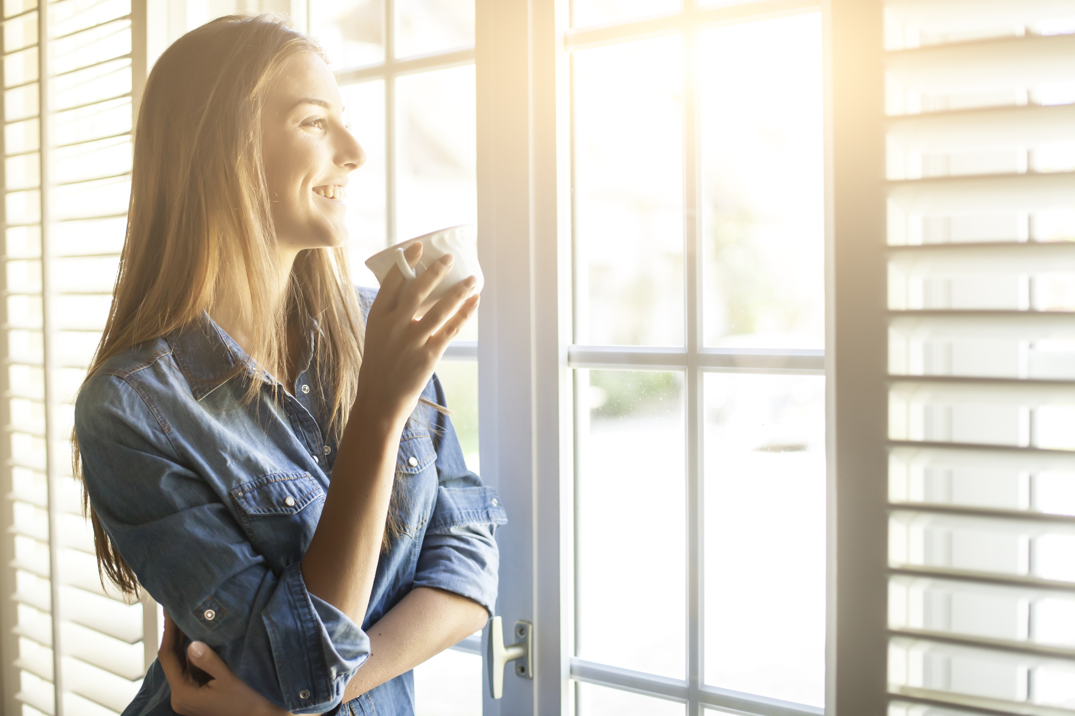 Young woman drinking coffee and looking through window as she considers roll-up shutters
